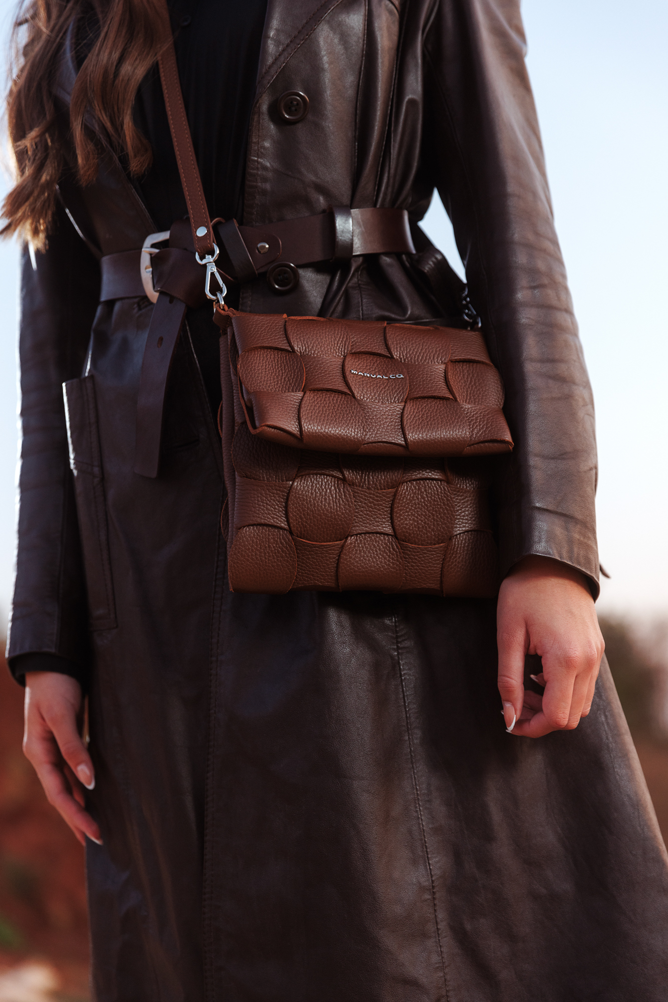 Person in brown leather trench coat carrying woven brown handbag, autumn fashion vibe.