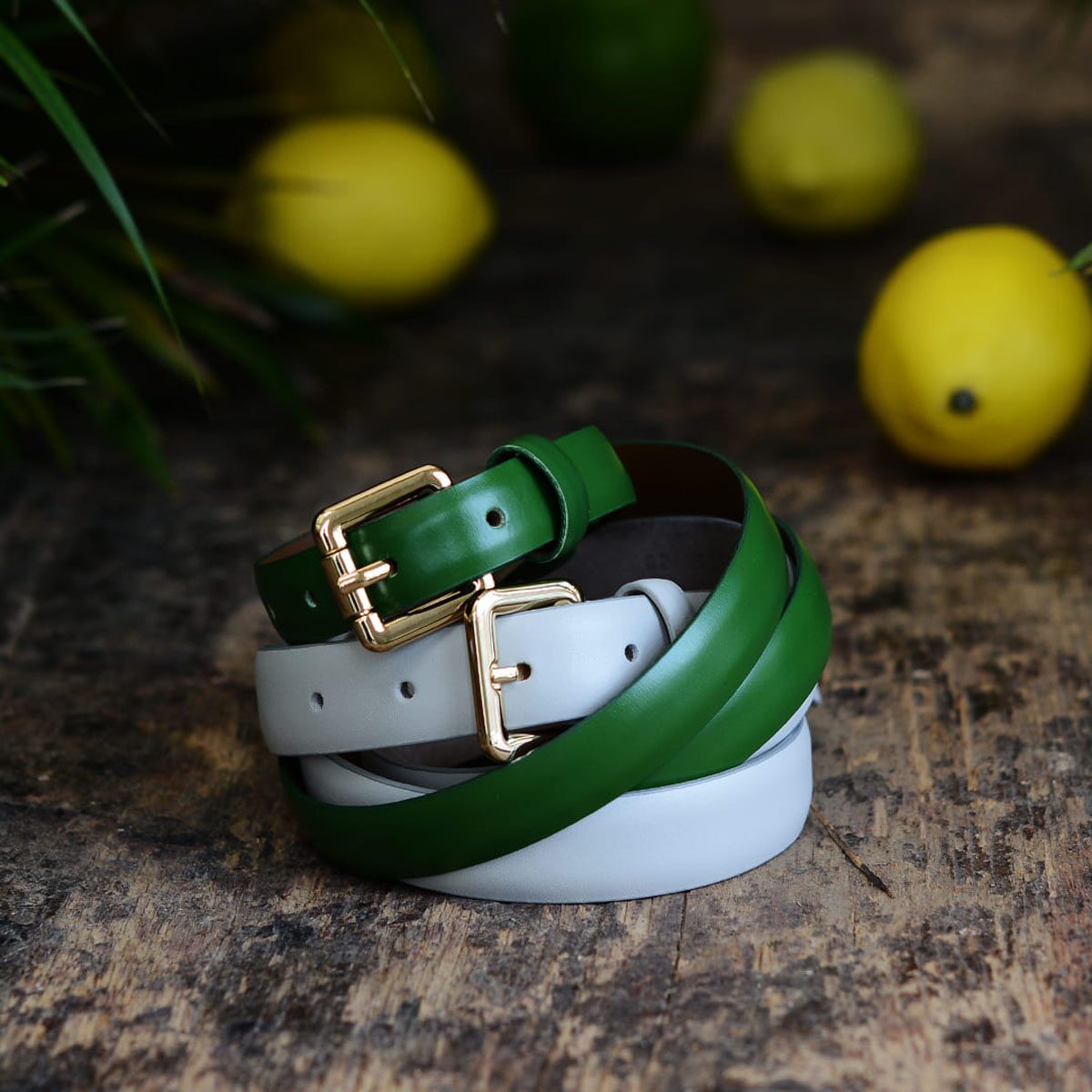 Green and white belts with gold buckles coiled on wood, citrus blurred in background.